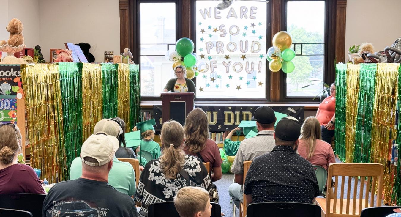 people sitting in chairs with a podium. Green and gold fringe curtains hang on the sides. 